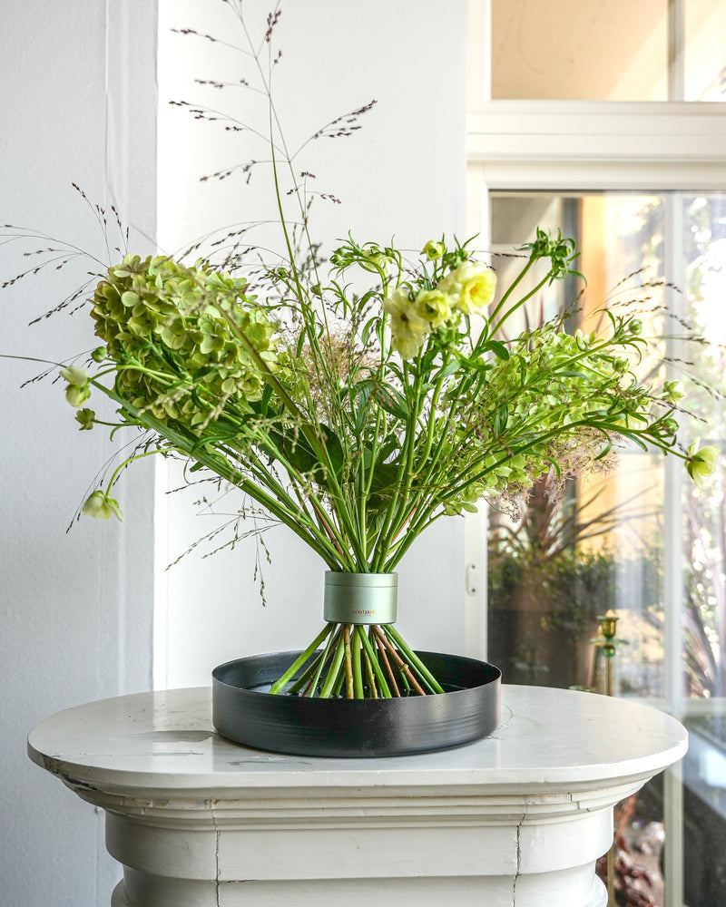 Bouquet professionale di grande volume in twister di fiori Hanataba Matcha Green, esposto su un vassoio floreale nero in cima a un piedistallo bianco.