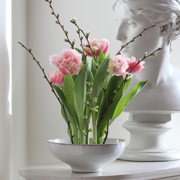 Elegante composizione floreale con tulipani rosa sfrangiati e delicati rami in boccio in una ciotola maculata, accostata a un busto scultura classico bianco.