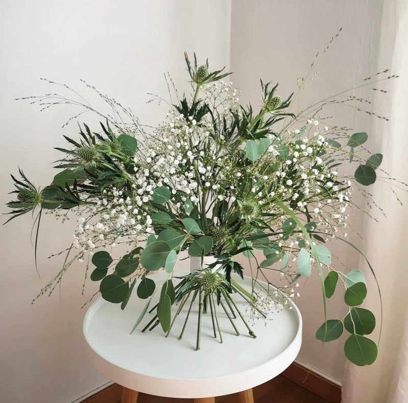 Elegante composizione ikebana con un assortimento di fiori verdi e bianchi, fissata da un Hanataba Bouquet Twister in argento perlato su un tavolo bianco.