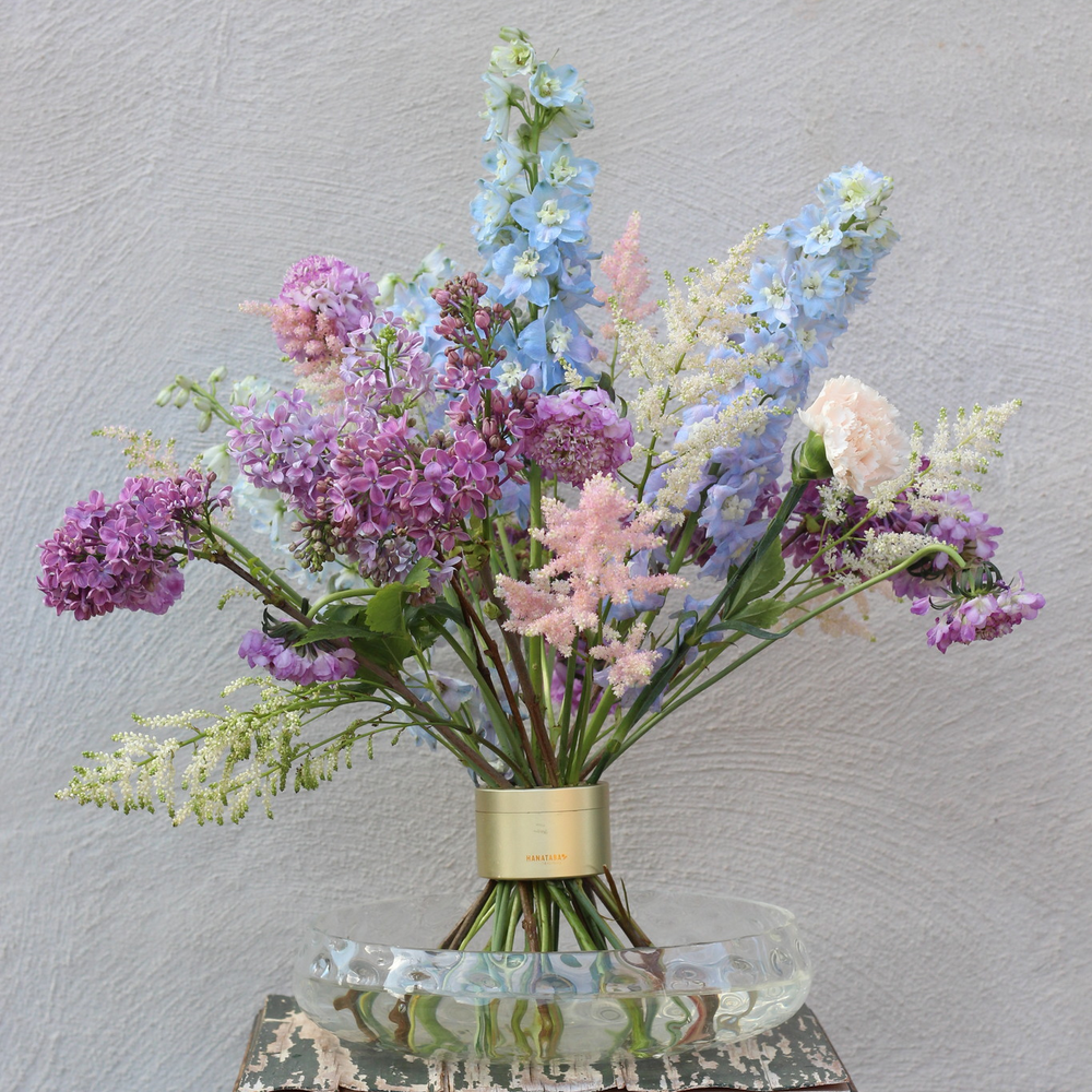 Elegante composizione floreale Ikebana con una varietà di fiori dai colori pastello, tra cui lillà e delphinium, fissati da un elegante supporto a stelo a spirale.