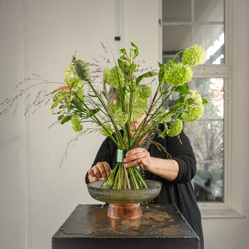 Un fiorista crea una composizione floreale professionale con fiori recisi di viburno in un verde edera Hanataba, esposti in un vassoio di vetro rotondo a righe.