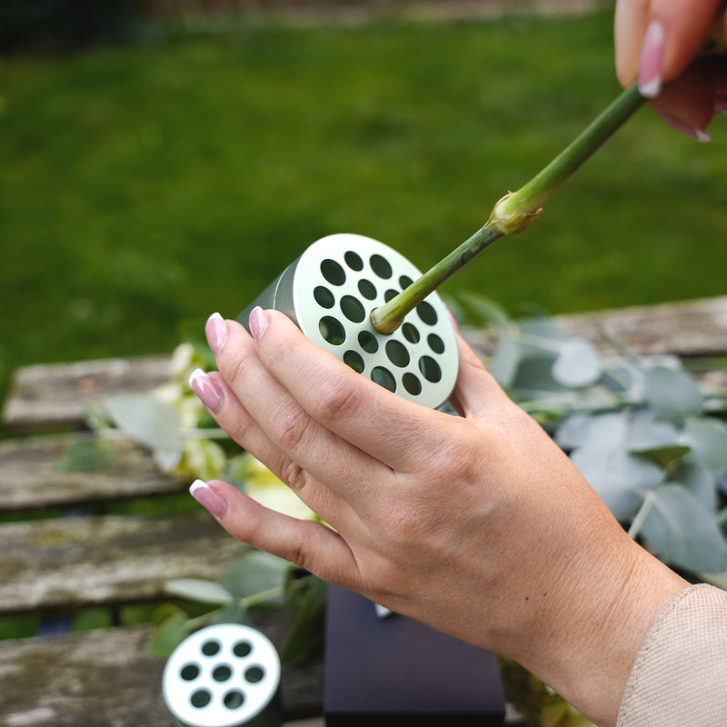 Una persona con lo smalto rosa che dispone eucalipti e fiori inserendo uno stelo in un attrezzo per la composizione di bouquet Hanataba Matcha Green su un tavolo di legno in giardino.