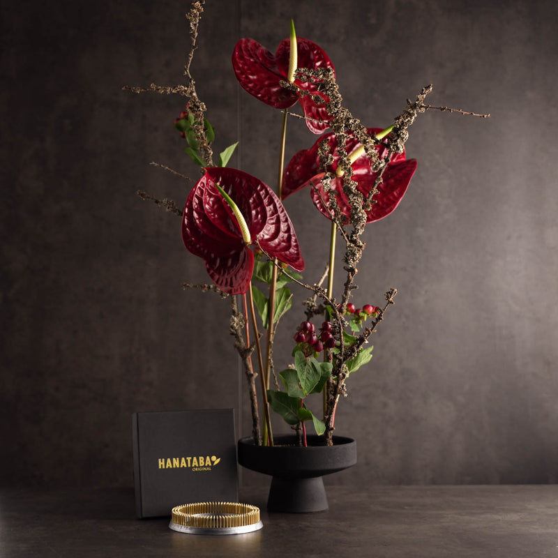 A modern kenzan floral arrangement of deep red anthurium flowers and branches in a black vase against a dark background. A small black box with "Hanataba" in gold lettering and round golden object are placed in front.