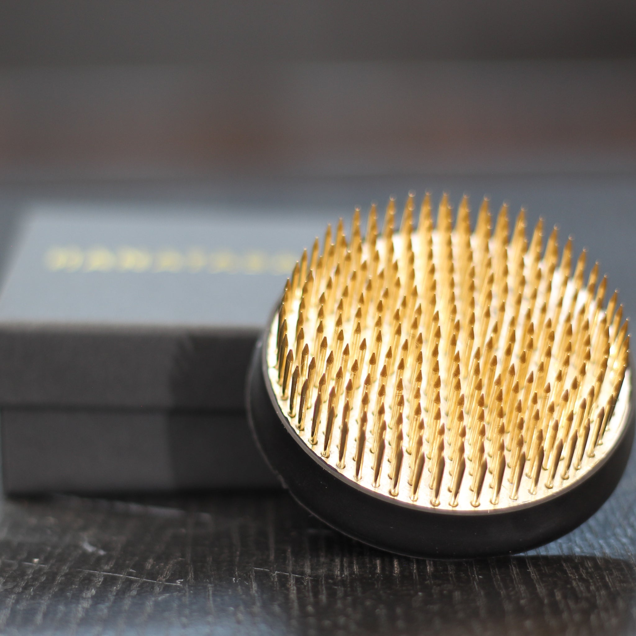 A small 70mm gold kenzan flower holder rests on a black base, with a blurred grey box featuring Japanese text in the background.