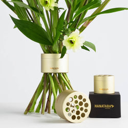 A beautifully designed bouquet of green-stemmed flowers sits elegantly in a circular, gold-colored Hanataba flower twister. Nearby, two similar Hanatabas are placed—one upright on a black packaging box and the other on its side, showcasing the unique circular pattern of holes.