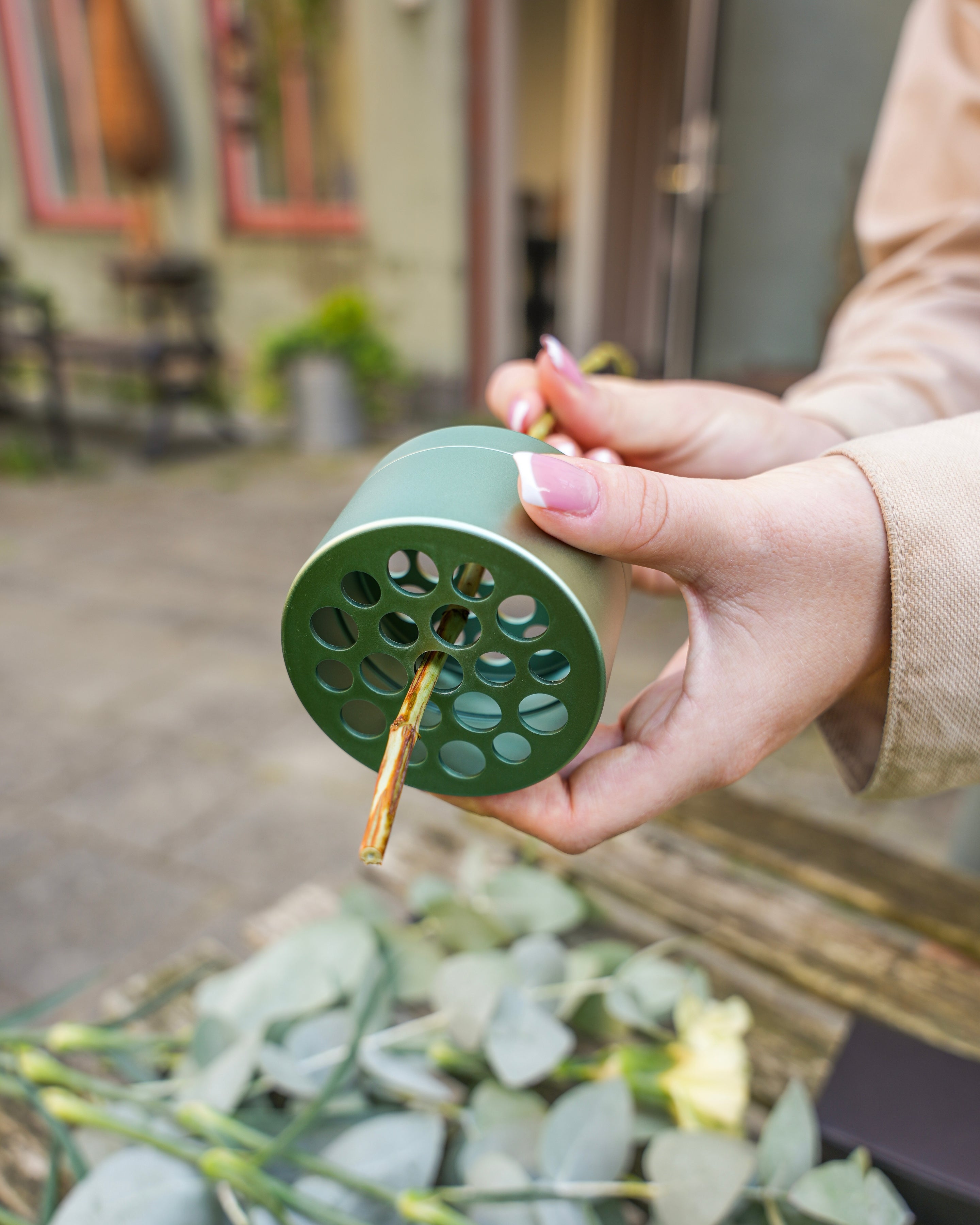 A person holding an Hanataba Ivy Green floral frog with a flower stem inserted through one of its holes. Eucalyptus leaves and other greenery are scattered on the surface below. The background shows an outdoor setting.