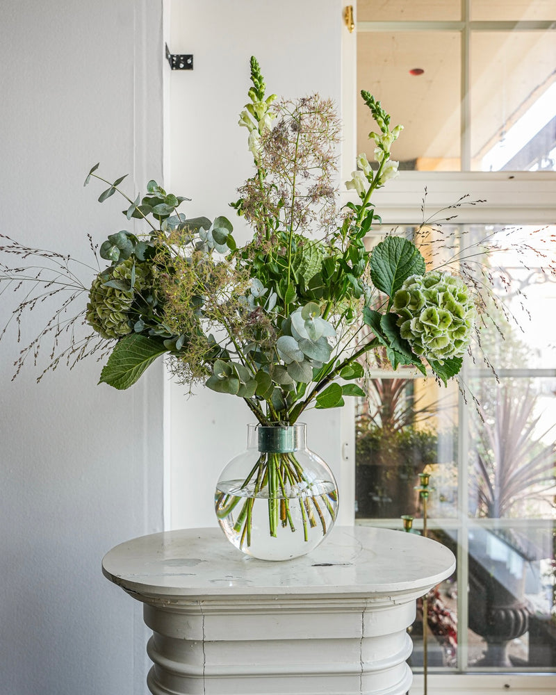 Un vaso di vetro trasparente con una composizione di fiori verdi e bianchi, tra cui ortensie ed eucalipti, si trova su un piccolo tavolo rotondo bianco. Il bouquet Hanataba Ivy Green spicca mentre la luce del sole filtra da un'alta finestra sullo sfondo.