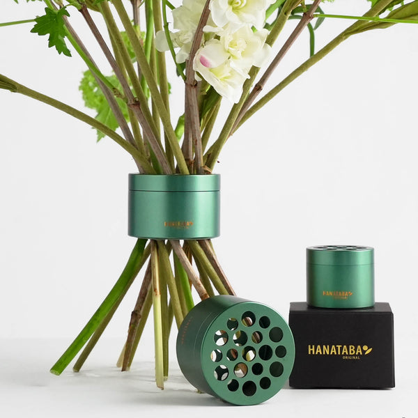 A bouquet of green stems with white flowers arranged with an Ivy Green Hanataba holder, inspired by ikebana floral design. Two additional Ivy Green holders are displayed: one standing on a black box with a gold Hanataba logo, another lying beside it, adding a refined touch to the floral display.”