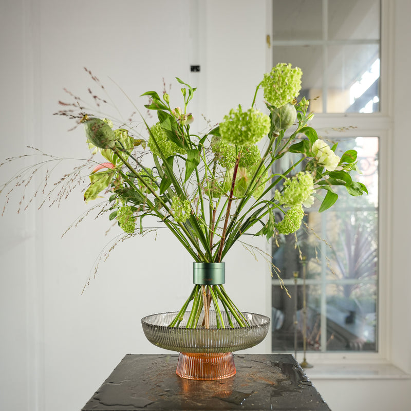 Splendido bouquet di fiori di graminacee e di viburno, disposti in uno strumento Hanataba Ivy Green, esposto in un vassoio di vetro con acqua su un tavolo nero vicino a una finestra in una stanza bianca e luminosa.