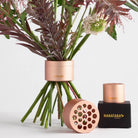 Pink metal ikebana-inspired floral arrangement tool with three cylindrical pieces, one showing the unique Hanataba product's stem-holding holes, positioned beside flowers. Another set rests on a black box labeled HANATABA in gold, enhancing a sophisticated bouquet display against a white background.