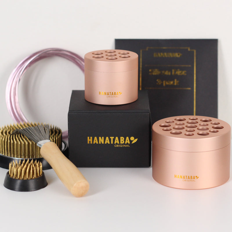 Un set di strumenti per la composizione floreale Hanataba Original, con due portafiori Hanataba Pink Champagne, un kenzan più grande (70 mm), un kenzan più piccolo (34 mm) e un pennello a rastrello kenzan con manico in legno. Gli articoli sono disposti su una scatola nera con scritte dorate del logo Hanataba.