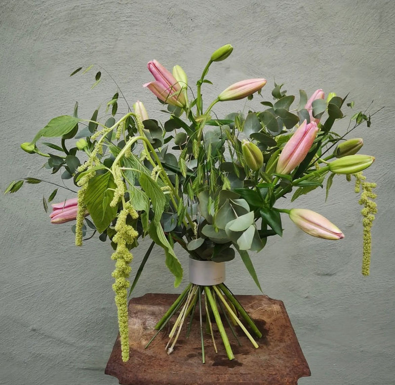 Una lussureggiante composizione floreale con gigli rosa e accenti verdi a cascata, il tutto tenuto insieme da un Hanataba Bouquet Twister in argento perlato, su una parete grigia strutturata.