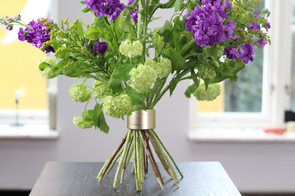 Bouquet vivace con fiori viola e viburno verde, disposto su un tavolo vicino a una finestra con un porta stelo a spirale Hanataba Original in oro champagne.