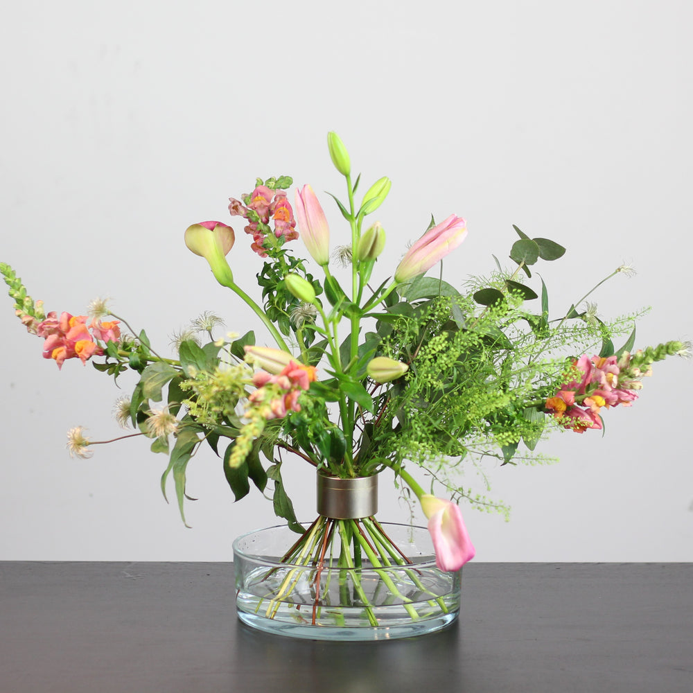 Un mix eterogeneo di fiori colorati disposti in un vaso a ciotola trasparente, tenuti insieme da un Hanataba Pitch Black Bouquet Twister