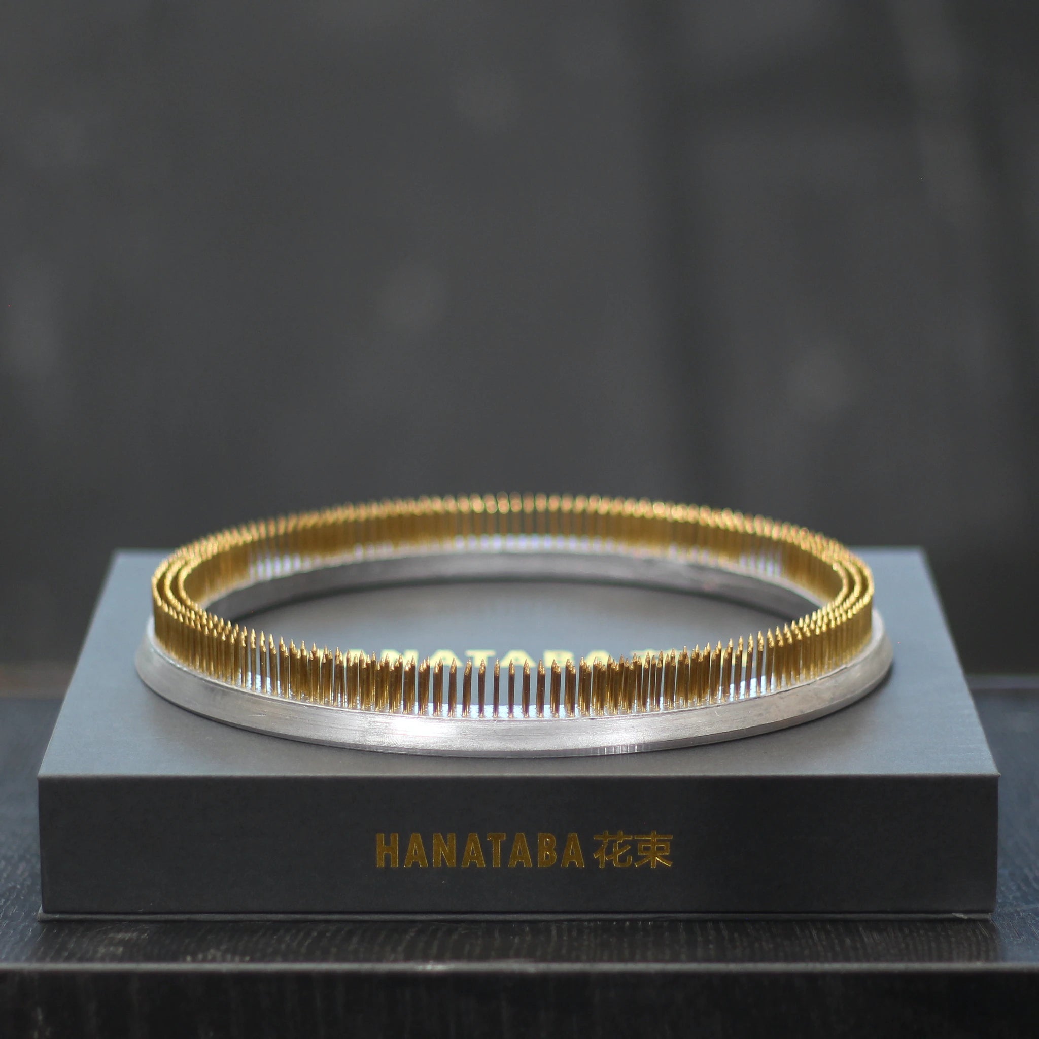 A gleaming 200mm Kenzan ring rests atop a sleek gray presentation box with the gold inscription "HANATABA 花束," showcasing elegance and functionality in floral arrangement.