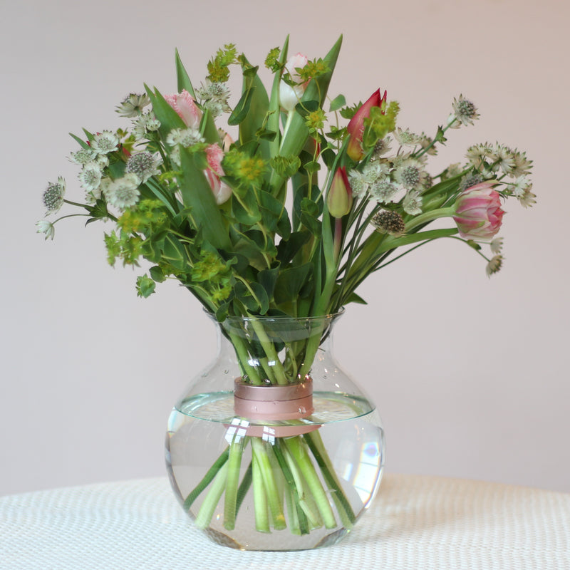 Un fresco bouquet di tulipani e piante verdi assortite, assicurato da un bouquet twister Hanataba rosa perlato, si trova in un vaso rotondo trasparente, che offre una miscela di eleganza e fascino primaverile.