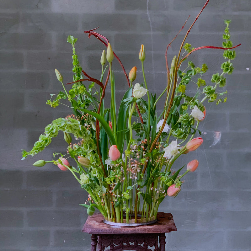 Un bouquet vibrante con tulipani e una serie di fiori verdi, meticolosamente disposti con un anello Kenzan da 200 mm su un piedistallo di legno ornato contro una parete di mattoni grigi.