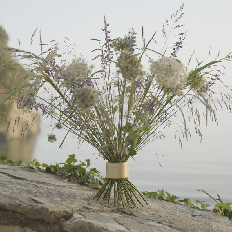 I fiori di campo, tra cui spighe viola e grappoli bianchi, si sono riuniti in un bouquet twister Hanataba color oro champagne su una sporgenza rocciosa che si affaccia su acque tranquille.