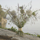 Fiori di campo ed erbe in un Hanataba Bouquet Twister Champagne Gold, in bilico su una roccia con l'acqua serena sullo sfondo, che trasmette una naturale tranquillità.